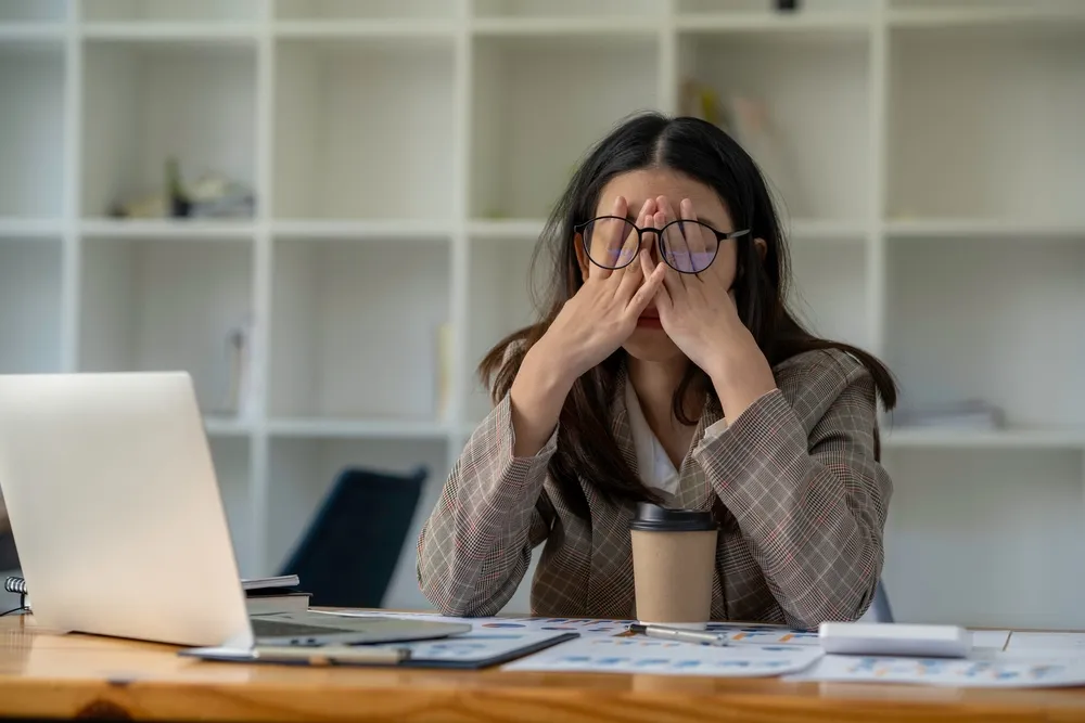 Una mujer con gafas sentada en un escritorio, frotándose los ojos con las manos, con un portátil, una taza de café y documentos en el escritorio, y estanterías blancas detrás.